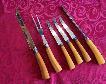 Op de afbeelding: Een snijset bestaande uit een groot mes, een vork met twee tanden, drie kleinere messen en een aanzetstaal. Elk stuk is voorzien van een gepolijst metalen lemmet of kop en een glad, lichtgeel handvat met een gebogen, ergonomische vorm.