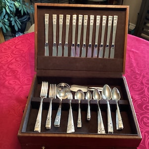 Puede incluir: Una caja de madera que contiene un juego de cubiertos. Los cubiertos están dispuestos en la caja, con los tenedores, los cuchillos y las cucharas en sus propios compartimentos. Los cubiertos son de plata y tienen un acabado pulido.
