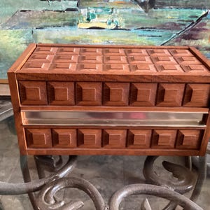 May include: A vintage wooden jewellery box with a geometric design on the lid and drawer fronts. The box is a warm brown colour with a silver-coloured metal band. The box is on a glass table.