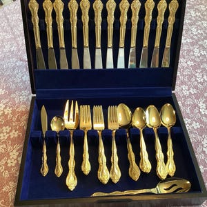 Può includere: Una scatola di legno con un interno in velluto blu che contiene un set di posate placcate oro. Il set comprende forchette, cucchiai e coltelli.