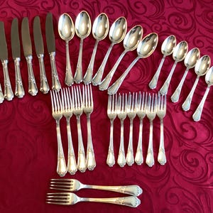 Puede incluir: Un conjunto de cubiertos plateados, que incluye cuchillos, cucharas y tenedores, dispuestos sobre un mantel rojo. La cubertería tiene mangos decorativos. El juego incluye varios tenedores, cucharas y cuchillos, adecuado para una cena formal.