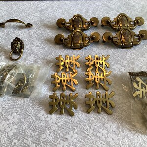 Puede incluir: Una colección de herrajes de latón antiguos, que incluyen tiradores de cajones, manijas y detalles decorativos. Los herrajes presentan diseños ornamentados y están empaquetados en plástico transparente. Las piezas están dispuestas sobre una superficie blanca con estampado floral.