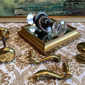 May include: A collection of door hardware including a crystal glass knob set with a dark metal base, two polished brass door knobs, and two curved brass door handles. The crystal knob features a circular metal plate with small embedded stones.