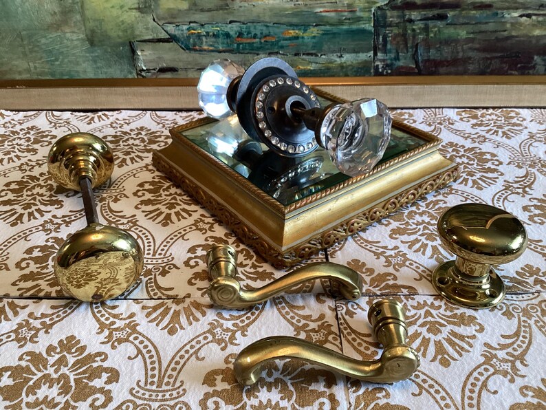 May include: A collection of door hardware including a crystal glass knob set with a dark metal base, two polished brass door knobs, and two curved brass door handles. The crystal knob features a circular metal plate with small embedded stones.