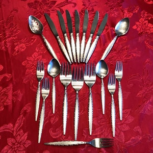 May include: A set of silverware with a decorative pattern on the handles. The set includes knives, forks, spoons, and a serving spoon. The silverware is arranged in a fan shape on a red tablecloth.