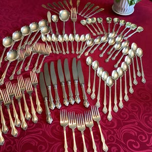 Könnte beinhalten: Eine Sammlung silberfarbener Besteckteile, darunter Gabeln, Messer und Löffel, auf einer roten Tischdecke angeordnet. Das Besteck wird in geordneter Weise präsentiert und zeigt verschiedene Teile für eine formelle Essumgebung. Die Anordnung deutet auf ein komplettes Set hin.