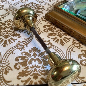 May include: A set of two polished brass door knobs connected by a square metal spindle. The knobs feature a smooth, rounded, reflective surface and are joined by a central rod, showcasing the hardware components used for door operation.
