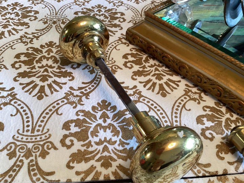 May include: A set of two polished brass door knobs connected by a square metal spindle. The knobs feature a smooth, rounded, reflective surface and are joined by a central rod, showcasing the hardware components used for door operation.