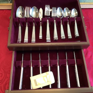 May include: A two-tiered wooden storage chest with a deep purple velvet interior containing a set of stainless steel cutlery. The top tray holds spoons, forks, and a knife, while the bottom drawer contains a row of knives and a small gold-toned rectangular metal tag.