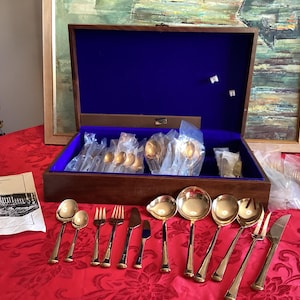 May include: A wooden silverware chest with a blue velvet interior holds gold-colored flatware. The set includes spoons, forks, knives, and serving pieces. The chest sits on a red tablecloth, with additional pieces displayed.