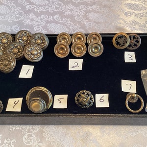 Può includere: Assortimento di 7 pomelli e maniglie per armadi in stile antico con finiture dorate e in ottone. I pomelli presentano disegni intricati, tra cui motivi floreali, geometrici e decorativi. Le maniglie sono di tipo ad anello e un pomello rotondo con un centro rialzato. I pomelli e le maniglie sono esposti su un vassoio di velluto nero con etichette numerate.