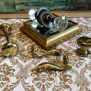 May include: A collection of door hardware including a crystal knob set with a dark metal base, two polished brass round knobs, and two curved brass door handles. The crystal knob set rests on a square gold-toned mirrored display base.