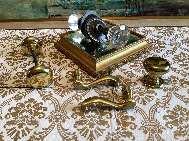 May include: A collection of door hardware including a crystal knob set with a dark metal base, two polished brass round knobs, and two curved brass door handles. The crystal knob set rests on a square gold-toned mirrored display base.