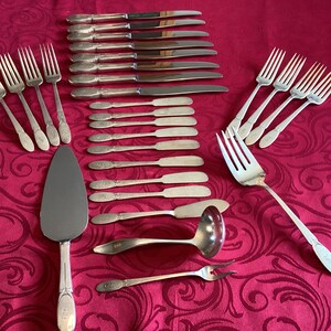 May include: A set of silverware, including forks, knives, and a serving spoon, all made of polished silver. The set is laid out on a red tablecloth with a floral pattern.