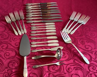 Può includere: Un set di posate, tra cui forchette, coltelli e un cucchiaio da portata, tutti in argento lucido. Il set è disposto su una tovaglia rossa con un motivo floreale.