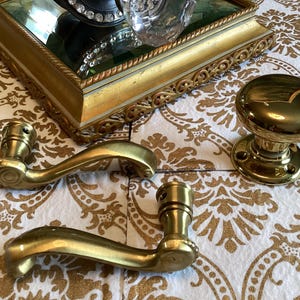 May include: A set of polished brass door hardware including two curved door handles with mounting points and one round door knob with a circular base plate. The hardware features a smooth, reflective metallic finish and classic, simple contours.