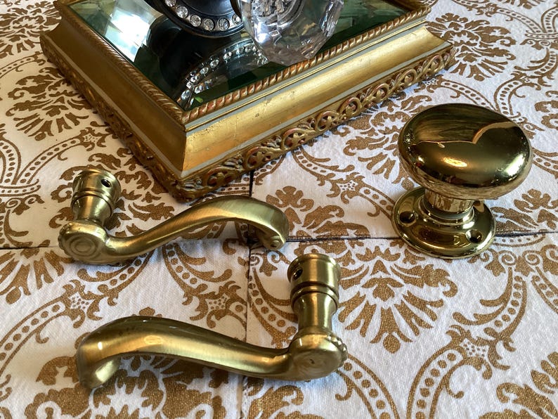 May include: A set of polished brass door hardware including two curved door handles with mounting points and one round door knob with a circular base plate. The hardware features a smooth, reflective metallic finish and classic, simple contours.