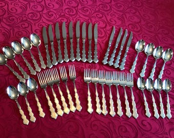 Könnte beinhalten: Ein komplettes Set aus Edelstahlbesteck, einschließlich Löffeln, Gabeln und Messern, angeordnet auf einer gemusterten roten Tischdecke. Das Besteck hat dekorative Griffe und eine polierte Oberfläche.