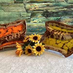 May include: Two decorative ceramic ashtrays with a brown rim and a wavy design. One is orange and brown, the other is yellow and brown. Sunflowers are placed between the ashtrays. The background is a painting with green and blue tones.