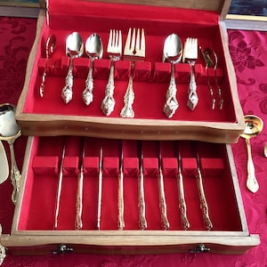 May include: A wooden cutlery chest with a red velvet interior, containing silver-coloured forks, spoons, and knives. Additional serving pieces are displayed outside the chest. The cutlery has ornate handles.