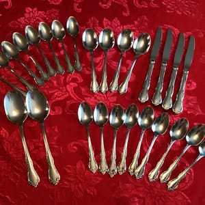 May include: A collection of silver-colored flatware, including spoons and knives, arranged on a red floral tablecloth. The spoons have a decorative handle design. The knives have a serrated edge.