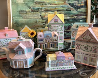 Op de afbeelding: Een verzameling keramische voorraadpotten in de vorm van huisjes en een theepot. De stukken hebben pastelkleuren, bloemmotieven en labels zoals Sugar, Utensils, Cookies, Rice, Village Tea Room en Butter. Elk item heeft een dakvormig deksel.