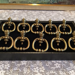 May include: Ten antique brass drawer pulls with a decorative design. The pulls are arranged in two rows of five in a black velvet lined box.