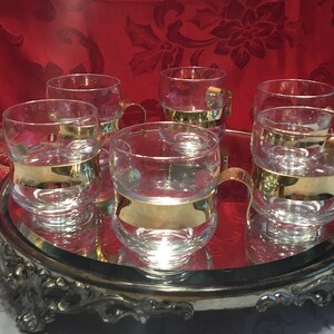May include: Set of six clear glass mugs with gold bands around the middle. The mugs are on a silver tray with a mirrored surface.