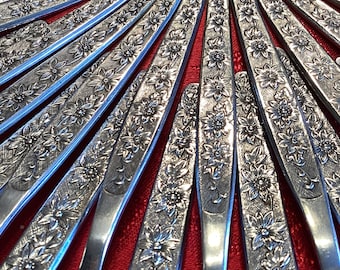 May include: A set of silver-plated knives with a floral design. The knives are arranged in a fan shape, with the handles pointing towards the center. The knives are all the same size and have a polished finish.