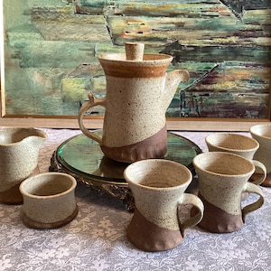 May include: A set of speckled brown and white ceramic mugs and a matching teapot with a lid. The mugs and teapot have a brown glaze on the bottom half. The set is on a white tablecloth with a floral pattern.