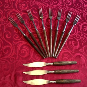 Könnte beinhalten: Ein Set Vintage-Besteck auf einer rot gemusterten Tischdecke. Das Set umfasst acht Gabeln mit silbernen Zinken und dunklen Holzgriffen sowie drei Buttermesser mit silbernen Klingen und passenden Griffen.