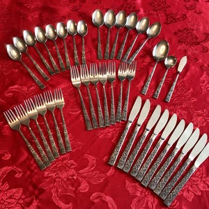 May include: A large collection of stainless steel flatware including dinner knives, dinner forks, salad forks, soup spoons, and teaspoons. Each piece features a textured, decorative handle design with a polished finish on the metal heads.
