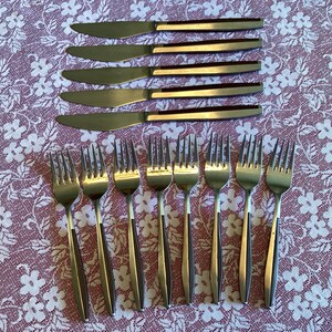 Könnte beinhalten: Ein Set aus Edelstahlbesteck, einschließlich Messer und Gabeln, angeordnet auf einer Tischdecke mit Blumenmuster. Die Messer haben eine goldfarbene Klinge und einen Griff, während die Gabeln silberfarben sind. Das Set enthält sechs Messer und acht Gabeln.