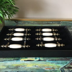 May include: A set of ten white ceramic cabinet knobs with brass accents. The knobs are displayed in a black velvet-lined box.