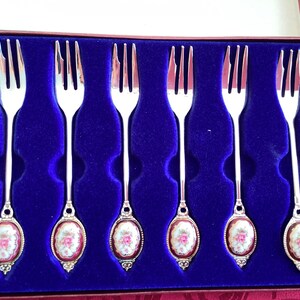 May include: Set of six silver dessert forks with porcelain handles featuring a floral design. The forks are displayed in a blue velvet lined box.
