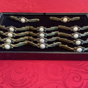 May include: A set of 12 antique-style cabinet knobs with a white stone inlay and a brass finish. The knobs are arranged in a box with black velvet lining.