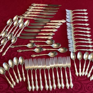 May include: A set of silverware with a silver finish. The set includes forks, knives, and spoons. The silverware is arranged in rows on a red tablecloth.