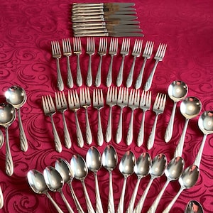 May include: A set of silverware with a silver finish. The set includes forks, spoons, and knives. The silverware is arranged in a fan shape on a red tablecloth.