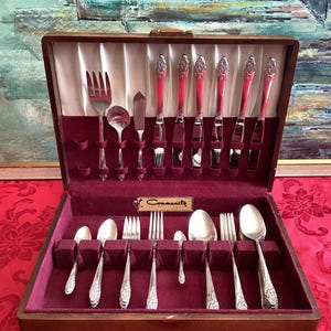 May include: A wooden silverware case with a burgundy velvet interior holds a complete set of Community silverware. The set includes forks, spoons, knives, and serving utensils. The silverware has ornate handles and a shiny finish, reflecting the light.