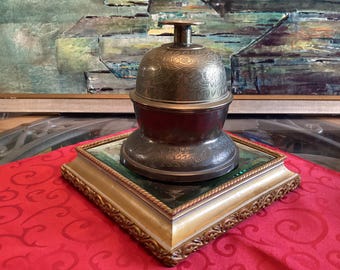 Op de afbeelding: Een metalen bureauklok met een ingewikkelde bloemengravure op de koepel en een plat handvat aan de bovenkant. De bel rust op een vierkante, goudkleurige voet met een decoratieve rand in touwstijl en een donker, reflecterend middenpaneel.