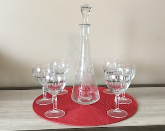 Op de afbeelding: Een set van zes heldere glazen wijnglazen met gegraveerde ontwerpen en een bijpassende karaf met stop. De glazen en de karaf zijn gerangschikt op een ronde rode placemat.