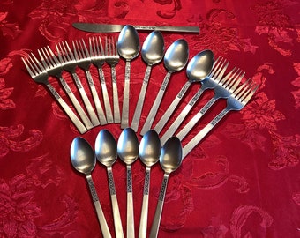 Puede incluir: Un juego de cubiertos, que incluye tenedores, cucharas y un cuchillo, con un patrón decorativo en los mangos. Los cubiertos están dispuestos en forma de abanico sobre un mantel rojo.