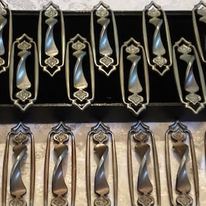 May include: A set of 12 antique silver-toned cabinet drawer pulls with a twisted design. The pulls are arranged in two rows of six, with the top row in a black velvet-lined box.