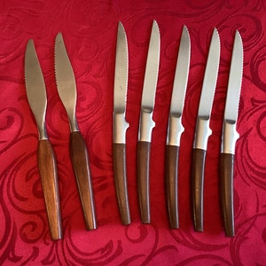 May include: Set of eight stainless steel steak knives with serrated blades and dark wood handles. The knives are arranged on a red patterned tablecloth, showcasing their polished metal and warm wood tones. A classic cutlery set.