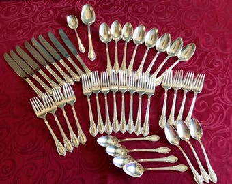 Op de afbeelding: Een grote set zilverkleurig roestvrijstalen bestek, inclusief dinermessen, dinervorken, saladevorken en lepels in verschillende maten. Elk stuk heeft een decoratief handvat met een subtiel reliëfpatroon aan de onderkant van de steel.