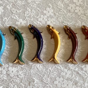 Puede incluir: Seis figuras de peces de cerámica coloridas en fila. Cada pez tiene un cuerpo curvo y una cola y detalles dorados. Los colores incluyen azul, verde, marino, amarillo, marrón y rojo. Las figuras están sobre una superficie blanca estampada.