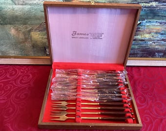 Op de afbeelding: Een houten presentatiedoos met een set goudkleurige metalen vorken met donkerbruine handvatten. De binnenkant van het deksel bevat de tekst James QUALITY JEWELLERS. Sommige vorken zijn verpakt in doorzichtig plastic, andere liggen bloot.