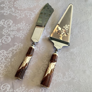 May include: A vintage cake serving set comprising a knife and server. Both have brown and cream carved handles and silver-toned blades. The server features a floral design.