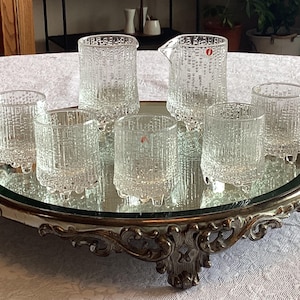 May include: A set of six clear glass drinking glasses with a textured pattern, including a pitcher, on a silver tray with an ornate design.