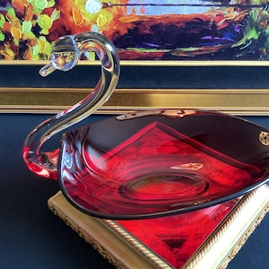 May include: A decorative glass swan-shaped bowl with a clear, curved neck and head. The bowl is a deep red color with a black rim and sits on a gold-colored stand. The background includes a framed painting.